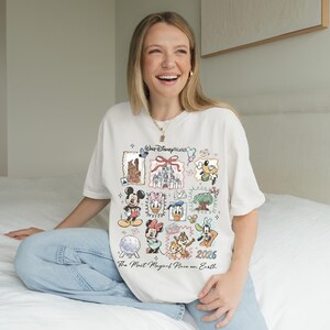 Puede incluir: Camiseta color crema con un dise&ntilde;o colorido de Disney World con varios personajes y lugares emblem&aacute;ticos. La camiseta incluye el texto "Walt Disney World" y "2026 The Most Magical Place on Earth."