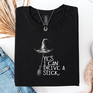 以下が含まれることがあります： 黒いTシャツに魔女の帽子とほうきのイラスト。テキスト「YES I CAN DRIVE A STICK」がグラフィックの下に印刷されています。太陽のペンダントが付いたシルバーのネックレスがシャツの上にあります。シャツは青いジーンズと白い靴と組み合わせています。