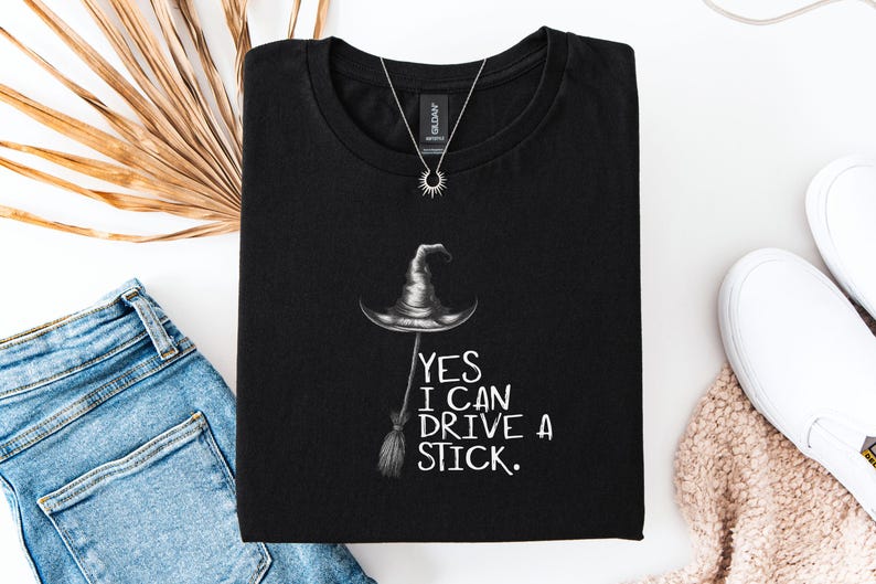 以下が含まれることがあります： 黒いTシャツに魔女の帽子とほうきのイラスト。テキスト「YES I CAN DRIVE A STICK」がグラフィックの下に印刷されています。太陽のペンダントが付いたシルバーのネックレスがシャツの上にあります。シャツは青いジーンズと白い靴と組み合わせています。