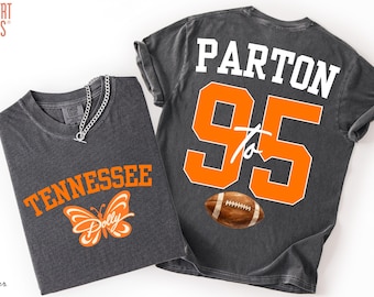 May include: Two charcoal grey t-shirts. The front features the orange text TENNESSEE above an orange butterfly with the word Dolly. The back displays the white text PARTON above the large orange number 95, the word to, and a brown football.