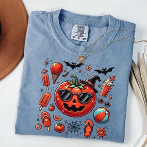 May include: A blue crew neck t-shirt featuring a graphic print of a smiling jack-o'-lantern wearing sunglasses and a witch hat, surrounded by summer-themed icons like ice cream, flip-flops, a beach ball, and bats, with a Comfort Colors label.