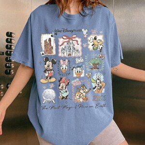 Puede incluir: Camiseta azul claro con personajes de Disney como Mickey, Minnie, Donald y Goofy. Incluye "Walt Disney World", "The Most Magical Place on Earth" y el a&ntilde;o "2026". El dise&ntilde;o presenta varias ilustraciones relacionadas con Disney.