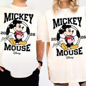 Puede incluir: Camisetas color crema con un gr&aacute;fico de Mickey Mouse. El dise&ntilde;o incluye a Mickey con uniforme de f&uacute;tbol, el texto "MICKEY MOUSE", el n&uacute;mero "26" y el logotipo de Disney. Las camisetas son de manga corta.