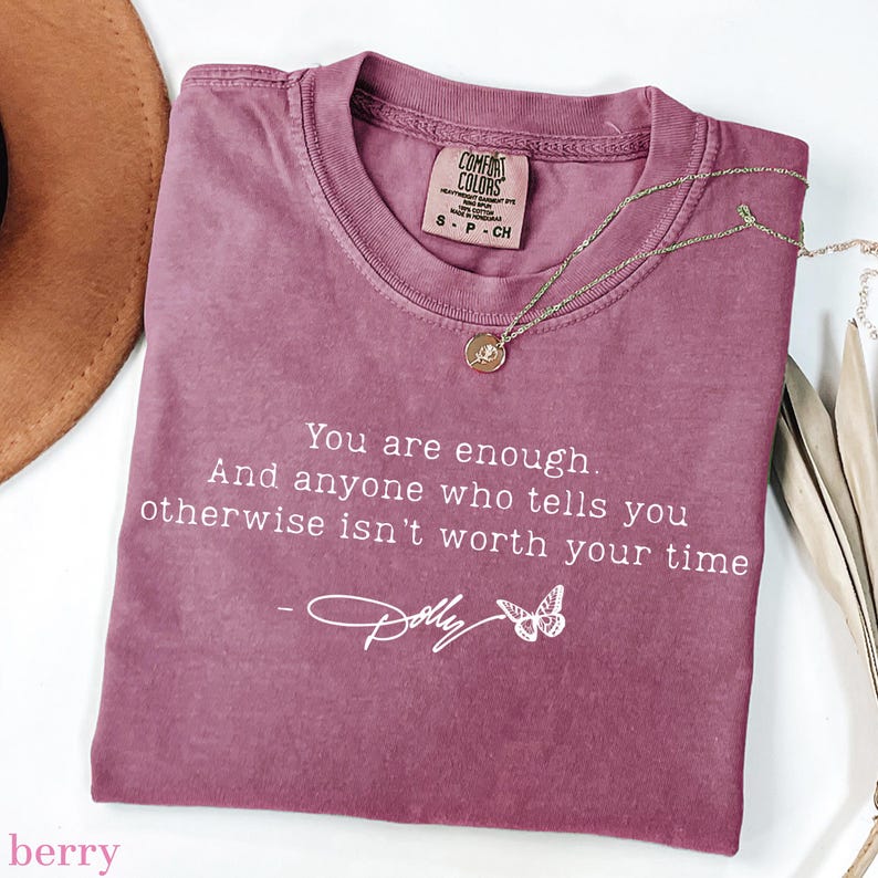May include: A berry-coloured short-sleeve t-shirt featuring the white printed text: You are enough. And anyone who tells you otherwise isn't worth your time - Dolly, accompanied by a small butterfly graphic. The shirt has a Comfort Colors brand label.