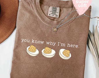 May include: Espresso-colored t-shirt with the phrase "you know why I'm here" and an illustration of three deviled eggs. A silver necklace with a pendant is draped across the shirt. The shirt has a "Comfort Colors" tag.
