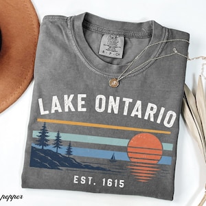 May include: A charcoal gray short-sleeve t-shirt featuring the text LAKE ONTARIO and EST. 1615 in white. The graphic includes a sunset, a sailboat, and pine trees on a shoreline in shades of orange, light blue, and dark blue.