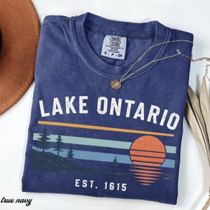 May include: A navy blue short-sleeve t-shirt featuring the white text LAKE ONTARIO and EST. 1615. The graphic includes a sunset over a lake with pine trees and a sailboat. A gold-toned necklace with a small circular pendant rests on the neckline.