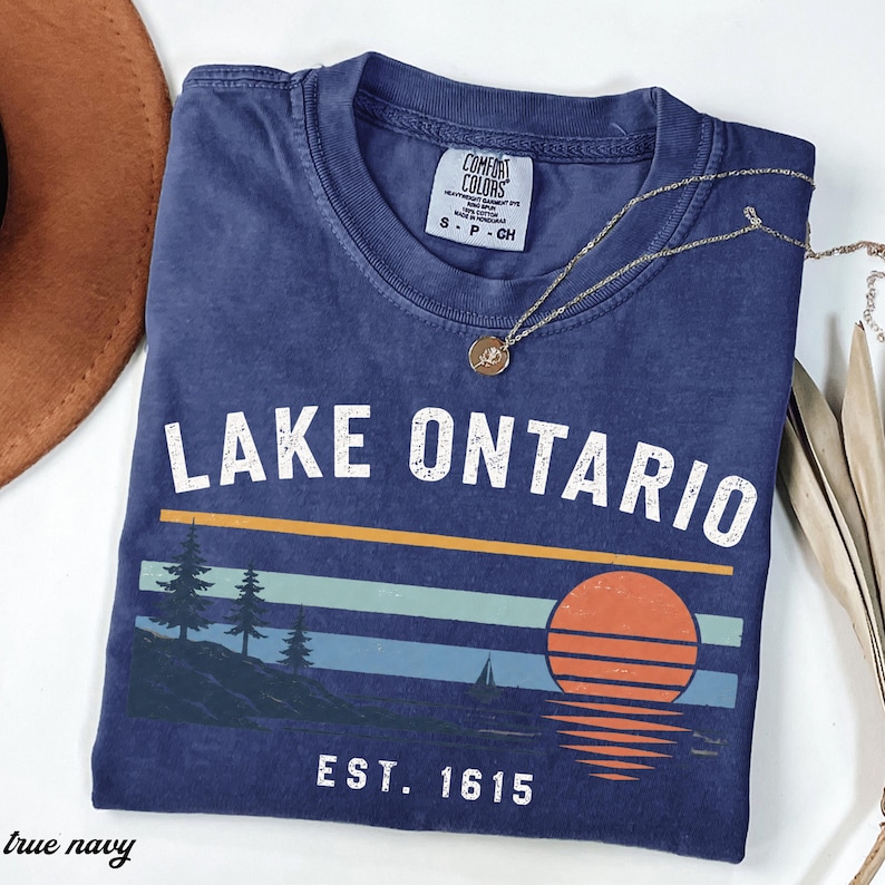 May include: A navy blue short-sleeve t-shirt featuring the white text LAKE ONTARIO and EST. 1615. The graphic includes a sunset over a lake with pine trees and a sailboat. A gold-toned necklace with a small circular pendant rests on the neckline.