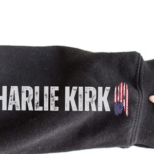 以下が含まれることがあります： 黒いスウェットシャツの袖に、使い古された白い文字で「CHARLIE KIRK」という名前が書かれています。袖にはアメリカ国旗のグラフィックがあります。画像は白い背景です。