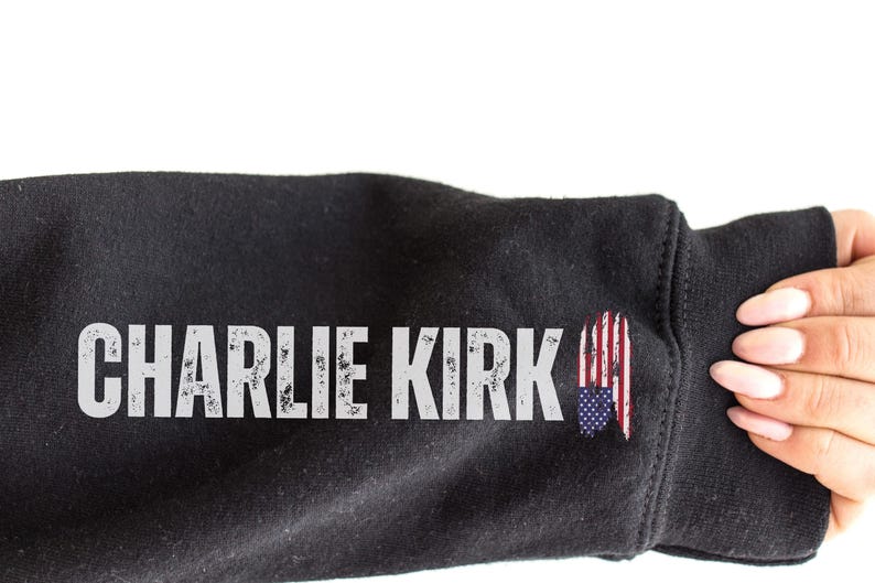 以下が含まれることがあります： 黒いスウェットシャツの袖に、使い古された白い文字で「CHARLIE KIRK」という名前が書かれています。袖にはアメリカ国旗のグラフィックがあります。画像は白い背景です。