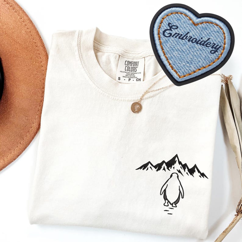 May include: Cream-colored t-shirt with a black penguin graphic walking towards a mountain range. A denim heart-shaped patch with the word "Embroidery" is visible. A gold necklace with a pendant is also present.