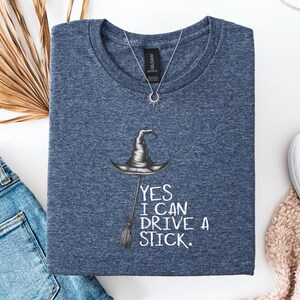 以下が含まれることがあります： 魔女の帽子とほうきのイラストが描かれたネイビーブルーのTシャツ。「YES I CAN DRIVE A STICK」の文字がプリントされています。太陽のペンダントが付いたシルバーのネックレスがTシャツの上にあります。