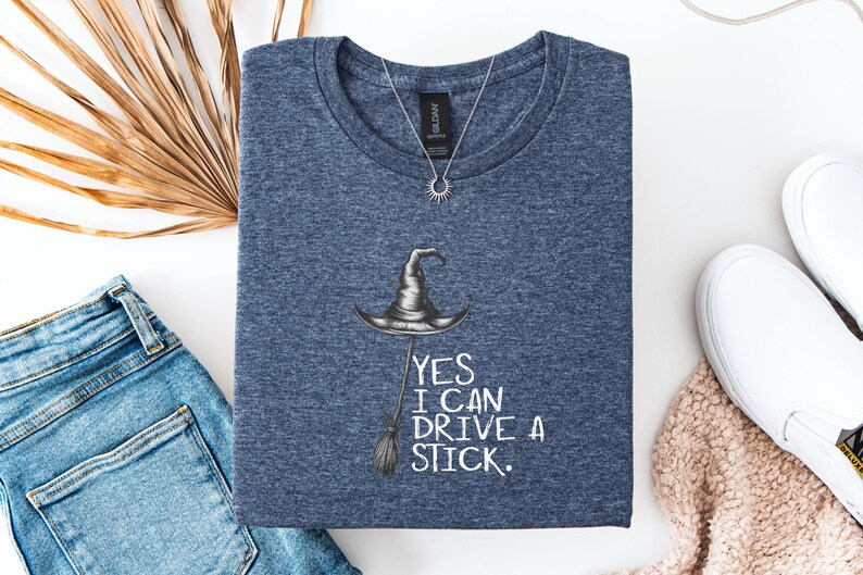 以下が含まれることがあります： 魔女の帽子とほうきのイラストが描かれたネイビーブルーのTシャツ。「YES I CAN DRIVE A STICK」の文字がプリントされています。太陽のペンダントが付いたシルバーのネックレスがTシャツの上にあります。