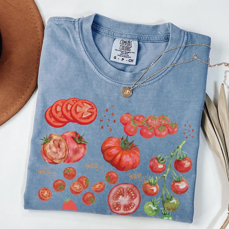 May include: A light blue t-shirt with a graphic print of red tomatoes. The shirt has the text "COMFORT COLORS" and "S - P - CH" printed on the tag.
