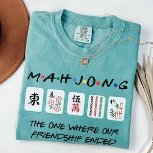 May include: A teal blue t-shirt with the words "Mahjong" and "The One Where Our Friendship Ended" printed on it. The shirt also features four mahjong tiles with Chinese characters.