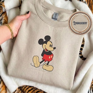 May include: A beige sweatshirt with an embroidered Mickey Mouse design. Mickey is shown in his classic outfit with red shorts, black top, and yellow shoes. The sweatshirt has a crew neck and a small tag.