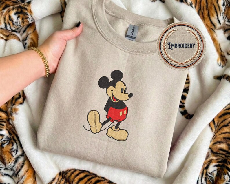 May include: A beige sweatshirt with an embroidered Mickey Mouse design. Mickey is shown in his classic outfit with red shorts, black top, and yellow shoes. The sweatshirt has a crew neck and a small tag.