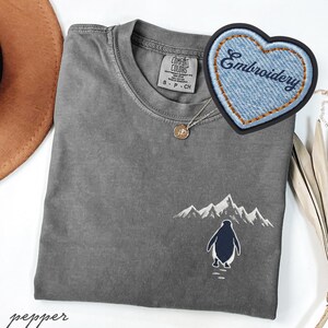 May include: A gray Comfort Colors t-shirt with an embroidered penguin and mountain scene. A denim heart-shaped patch with the word "Embroidery" is visible. A gold pendant necklace is also present.