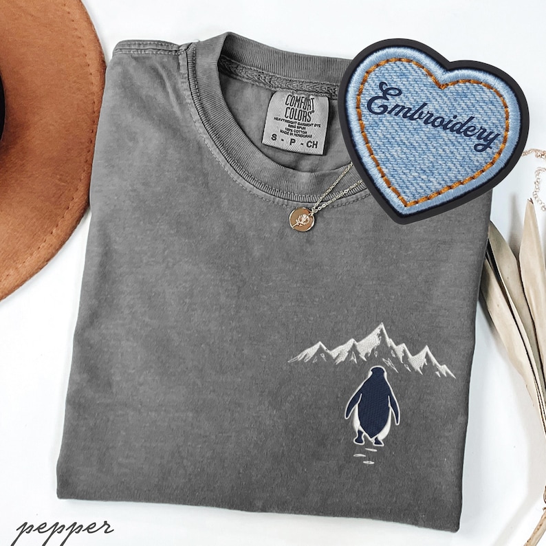 May include: A gray Comfort Colors t-shirt with an embroidered penguin and mountain scene. A denim heart-shaped patch with the word "Embroidery" is visible. A gold pendant necklace is also present.