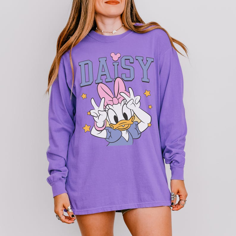 Puede incluir: Una camiseta morada de manga larga con un dise&ntilde;o de Daisy Duck, un lazo rosa y signos de paz. La palabra "DAISY" est&aacute; impresa en letras azules sobre el personaje, con peque&ntilde;as estrellas amarillas.