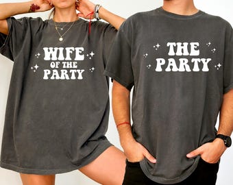 May include: Two charcoal gray t-shirts with white text. One shirt says "WIFE OF THE PARTY" and the other says "THE PARTY". Both shirts have a relaxed fit and are suitable for couples or groups.