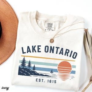 May include: An ivory t-shirt featuring a graphic of Lake Ontario with a sunset, pine trees, and a sailboat. The text reads Lake Ontario and Est. 1615. The shirt has a Comfort Colors label in the collar and is paired with a gold necklace and hat.
