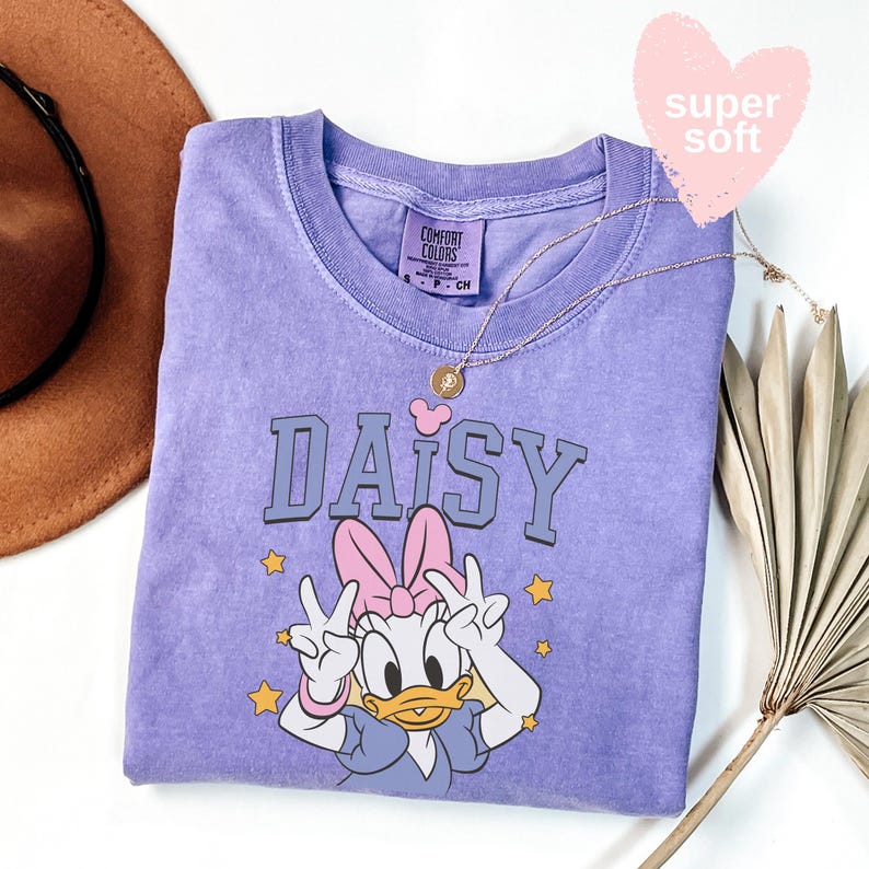 Puede incluir: Una camiseta lavanda Comfort Colors con un gr&aacute;fico de Daisy Duck y la palabra "DAISY" en azul. Daisy hace el signo de la paz. Tambi&eacute;n se ven un collar dorado y una etiqueta rosa en forma de coraz&oacute;n que dice "super soft".