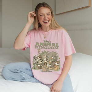 May include: A light pink t-shirt featuring the text "Disney's Animal Kingdom" in green, with a graphic of various Disney characters and animals. The shirt is a relaxed fit.