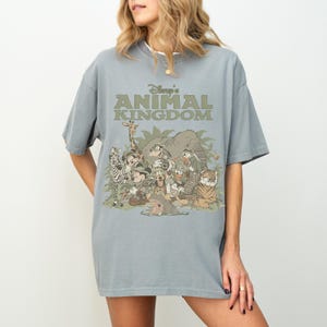 May include: A light blue t-shirt featuring the text "Disney's Animal Kingdom" in green. The graphic includes various cartoon animals such as a zebra, giraffe, elephant, and tiger. The shirt is a relaxed fit.