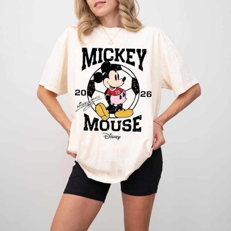 Puede incluir: Camiseta color crema con un dise&ntilde;o de Mickey Mouse con uniforme de f&uacute;tbol, con el texto "MICKEY MOUSE" arriba y abajo de la imagen. La camiseta incluye el logo de Disney y los n&uacute;meros "20" y "26".