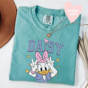 Puede incluir: Camiseta Comfort Colors de color verde azulado con un dise&ntilde;o de Daisy Duck, un lazo rosa y la palabra "DAISY" en morado. La camiseta tiene cuello redondo y mangas cortas. Tambi&eacute;n se ve un collar dorado y una pegatina rosa en forma de coraz&oacute;n.