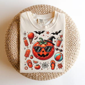 May include: A cream-colored t-shirt featuring a central graphic of a jack-o'-lantern wearing sunglasses and a witch hat, surrounded by summer-themed icons like ice cream, popsicles, flip-flops, a beach ball, bats, and a spider web.