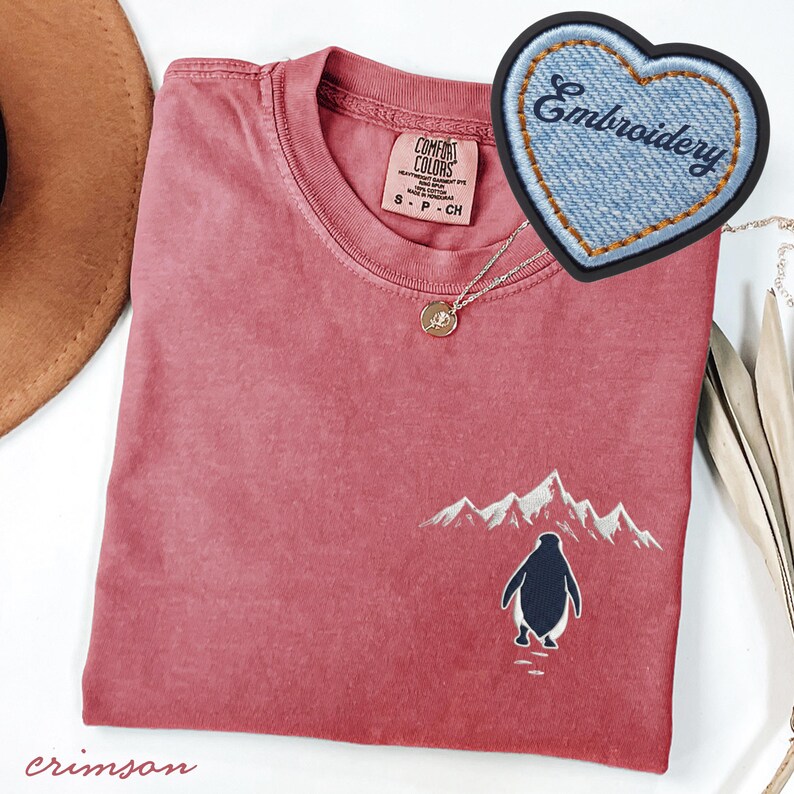 May include: Crimson-colored t-shirt with a crew neck and short sleeves. The shirt features an embroidered design of a penguin walking towards snow-capped mountains. A denim heart-shaped patch with the word "Embroidery" is visible.
