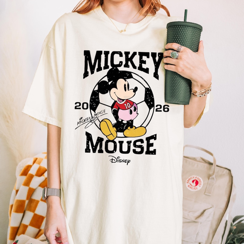 Puede incluir: Camiseta color crema con un gr&aacute;fico de Mickey Mouse dentro de un bal&oacute;n de f&uacute;tbol. El dise&ntilde;o incluye el texto "MICKEY MOUSE" y "20 26". El logotipo de Disney est&aacute; en la parte inferior. La camiseta es de corte holgado.