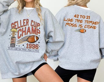 Op de afbeelding: Een lichtgrijs sweatshirt met de tekst "GELLER CUP CHAMP 1996" en een voetbalafbeelding. De achterkant van het sweatshirt heeft de tekst "42 TO 21 LIKE THE TURKEY ROSS IS DONE" en een voetbalafbeelding.