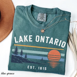 May include: A blue-green short-sleeve t-shirt featuring the text LAKE ONTARIO and EST. 1615 in white. The graphic includes a sunset, a sailboat, and pine trees on a shoreline. The shirt has a Comfort Colors label and a crew neckline.