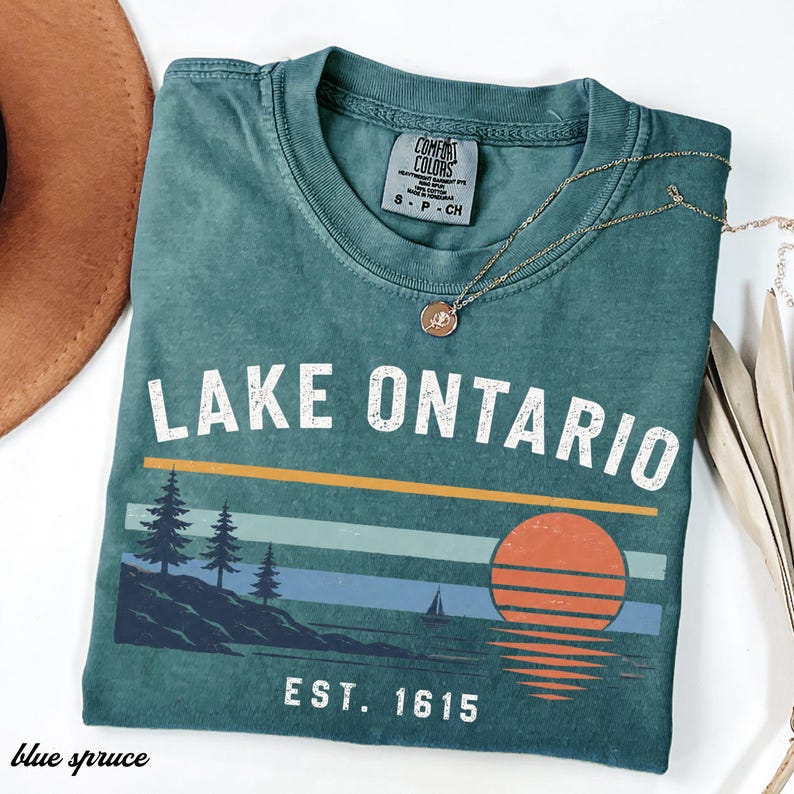 May include: A blue-green short-sleeve t-shirt featuring the text LAKE ONTARIO and EST. 1615 in white. The graphic includes a sunset, a sailboat, and pine trees on a shoreline. The shirt has a Comfort Colors label and a crew neckline.