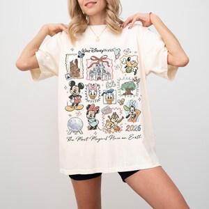Puede incluir: Camiseta de color crema con un gr&aacute;fico colorido de varios personajes y lugares emblem&aacute;ticos de Disney. El dise&ntilde;o incluye a Mickey Mouse, Minnie Mouse, el Pato Donald y el castillo de Cenicienta. El texto en la camiseta dice "Walt Disney World" y "The Most Magical Place on Earth" con el a&ntilde;o "2026".