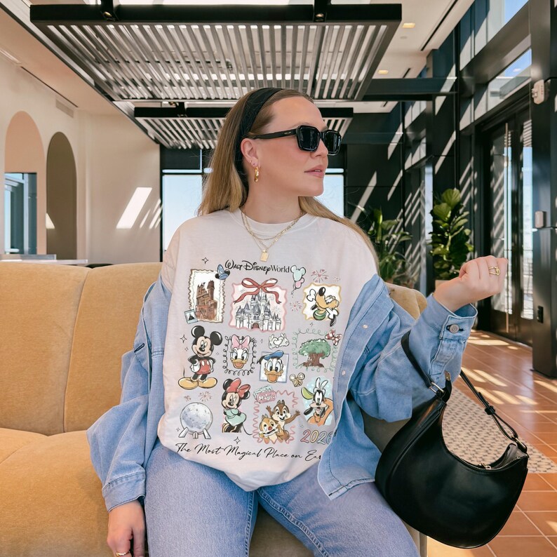 Puede incluir: Una camiseta color crema con varios personajes y lugares emblem&aacute;ticos de Disney, como Mickey Mouse, Minnie Mouse y el castillo de Cenicienta. La camiseta tiene el texto "Walt Disney World" y "The Most Magical Place on Earth."