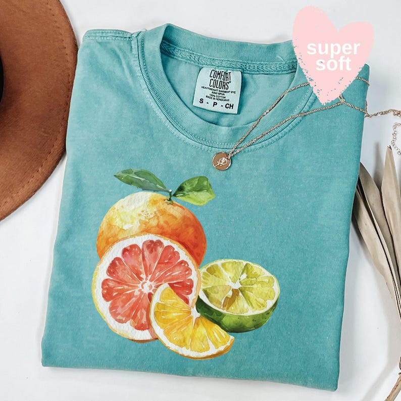 Op de afbeelding: Een turquoise Comfort Colors t-shirt met een aquarel illustratie van citrusvruchten. Het ontwerp bevat een hele sinaasappel, een halve grapefruit en limoen- en sinaasappelschijfjes. Een gouden ketting met een hanger is over het shirt gedrapeerd.