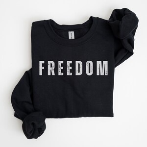 以下が含まれることがあります： 胸に白いブロック体で「FREEDOM」の文字がプリントされた黒色の長袖スウェットシャツ。スウェットシャツは白い面に折りたたまれています。袖は両側に折りたたまれています。