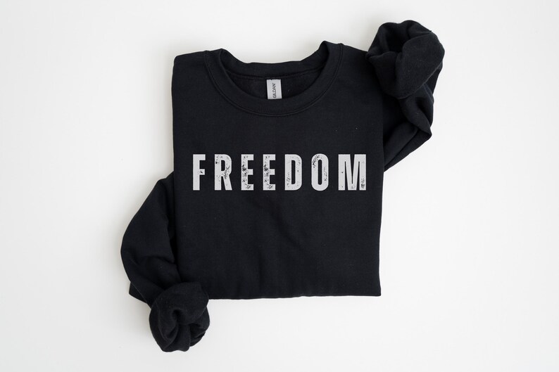 以下が含まれることがあります： 胸に白いブロック体で「FREEDOM」の文字がプリントされた黒色の長袖スウェットシャツ。スウェットシャツは白い面に折りたたまれています。袖は両側に折りたたまれています。