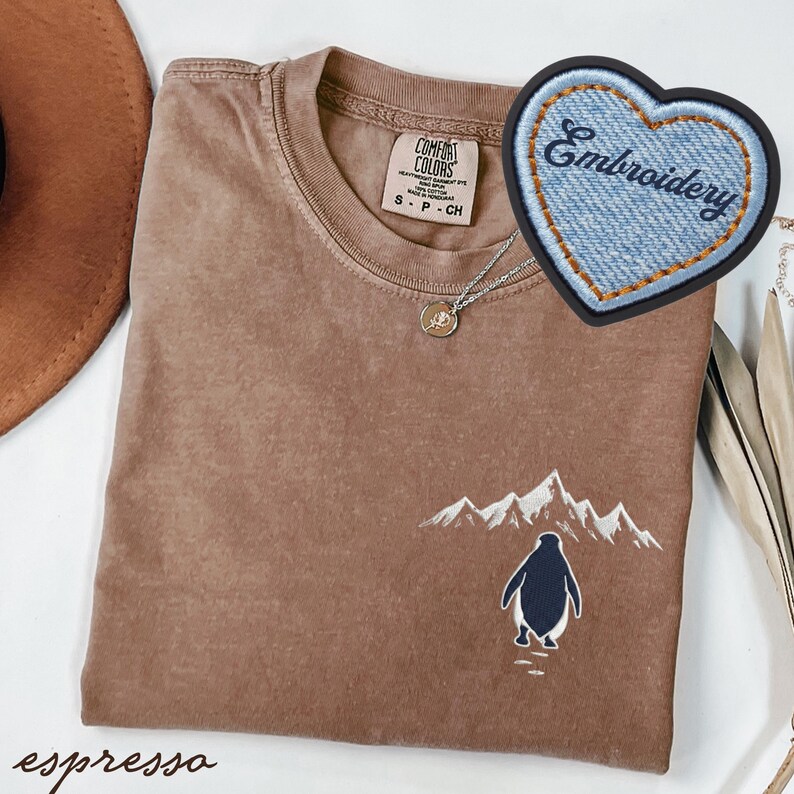 May include: Espresso-colored t-shirt with an embroidered penguin and mountain scene. Features a Comfort Colors tag and a denim heart patch with "Embroidery" text. A silver necklace with a pendant is also visible.