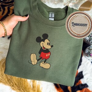 May include: An olive green sweatshirt with an embroidered Mickey Mouse design. The cartoon character is shown in his classic outfit with red shorts and tan shoes. A circular patch with the word "EMBROIDERY" is also visible.