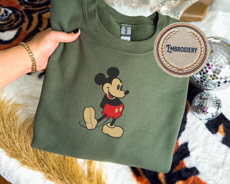 May include: An olive green sweatshirt with an embroidered Mickey Mouse design. The cartoon character is shown in his classic outfit with red shorts and tan shoes. A circular patch with the word "EMBROIDERY" is also visible.