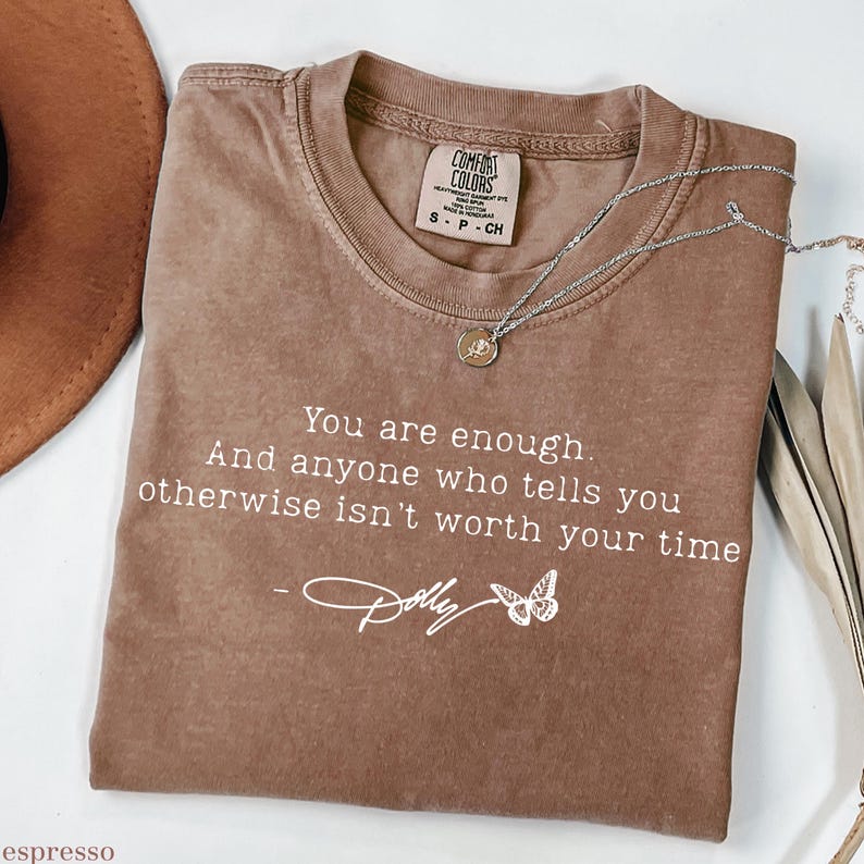 May include: A brown short-sleeved t-shirt with a crew neckline. The shirt features white text that reads: You are enough. And anyone who tells you otherwise isn't worth your time - Dolly, accompanied by a small white butterfly graphic.