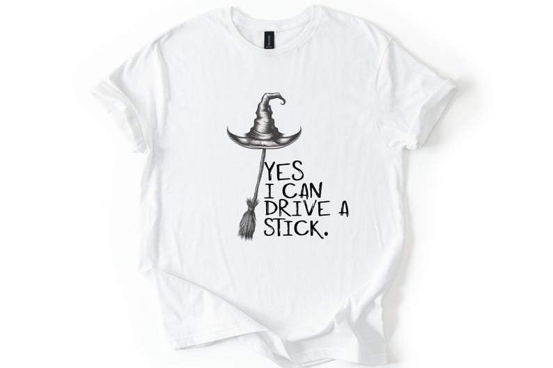 以下が含まれることがあります： 白のTシャツ。黒とグレーの魔女の帽子とほうきのイラスト。イラストの下には「YES I CAN DRIVE A STICK」の文字が黒でプリントされています。