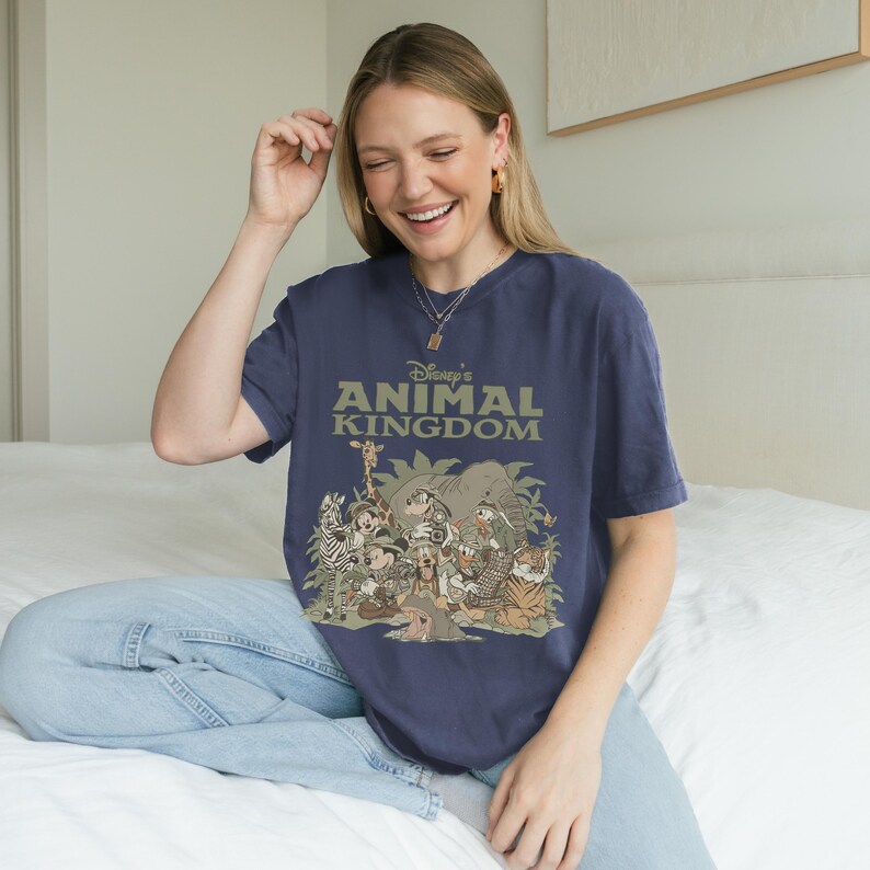 May include: A navy blue t-shirt featuring the text "Disney's Animal Kingdom" in green. The shirt has a graphic of various cartoon animals, including a giraffe, zebra, elephant, and tiger. The shirt is made of a soft material.