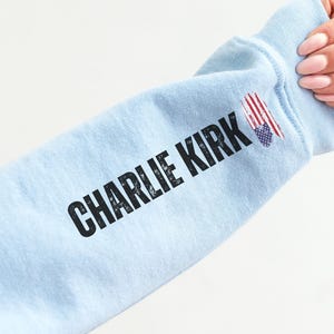 以下が含まれることがあります： 黒い文字で「CHARLIE KIRK」と書かれた、水色のスウェットシャツの袖。袖には小さなアメリカ国旗のグラフィックがあります。袖は人が持っています。