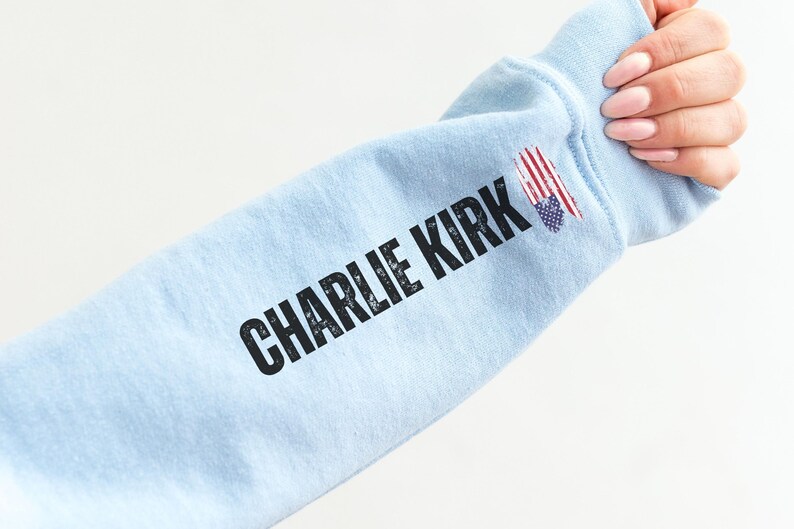 以下が含まれることがあります： 黒い文字で「CHARLIE KIRK」と書かれた、水色のスウェットシャツの袖。袖には小さなアメリカ国旗のグラフィックがあります。袖は人が持っています。